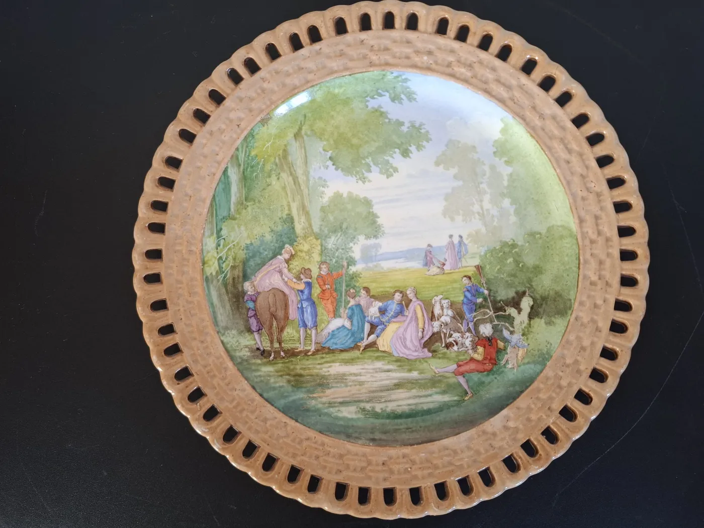 Assiette au bord ajouré, peinte d'une scène galante dans un parc : des personnages en costumes d'époque, un cheval et des chiens.