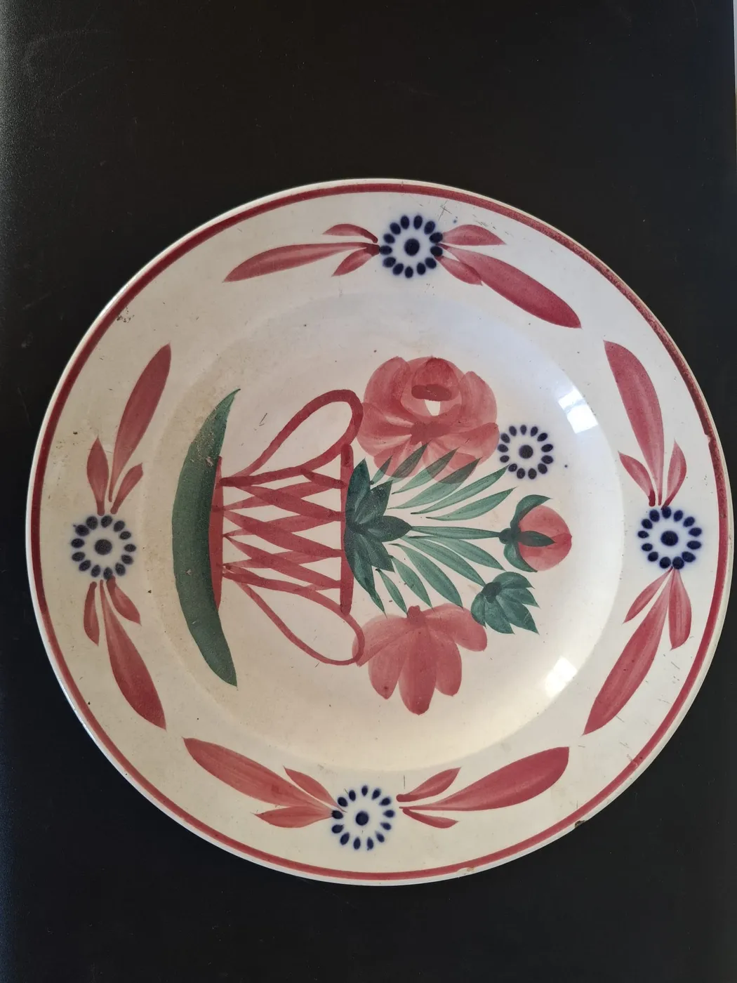 Assiette en faïence peinte à la main : un panier de fleurs roses et de feuillage vert au centre, une frise de feuilles roses et de fleurs bleues sur le bord.