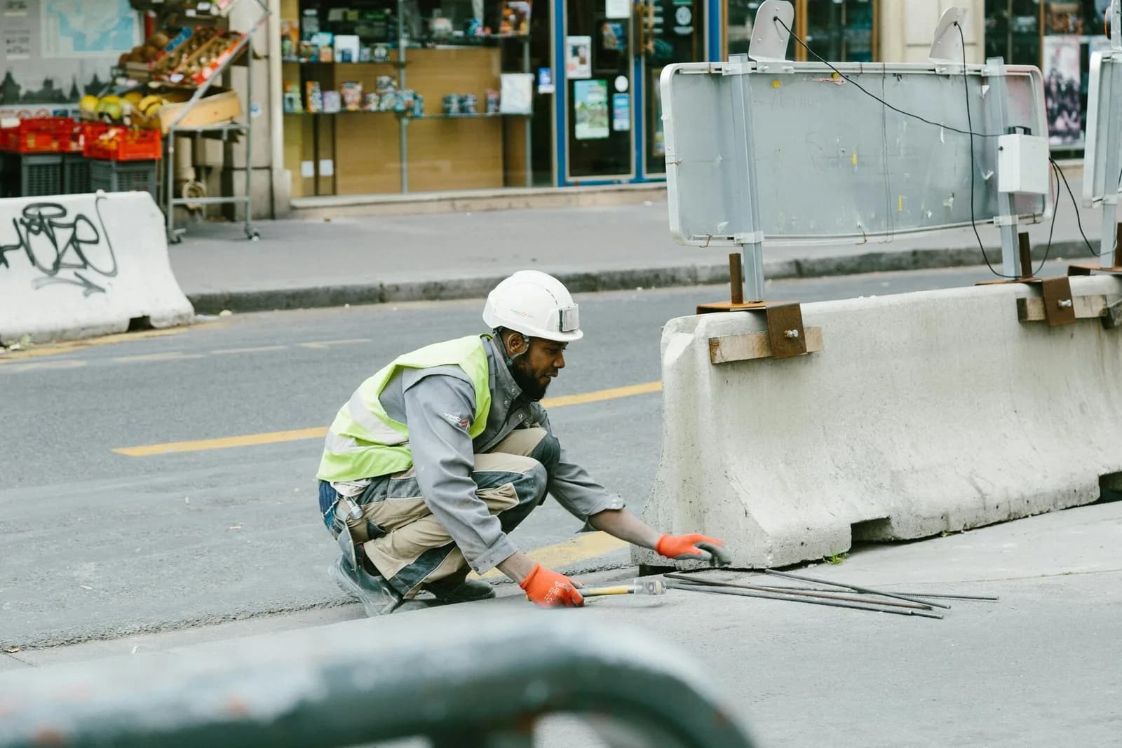 Un ouvrier en casque et gilet jaune accroupi au bord d'une route, près de blocs de béton de chantier.