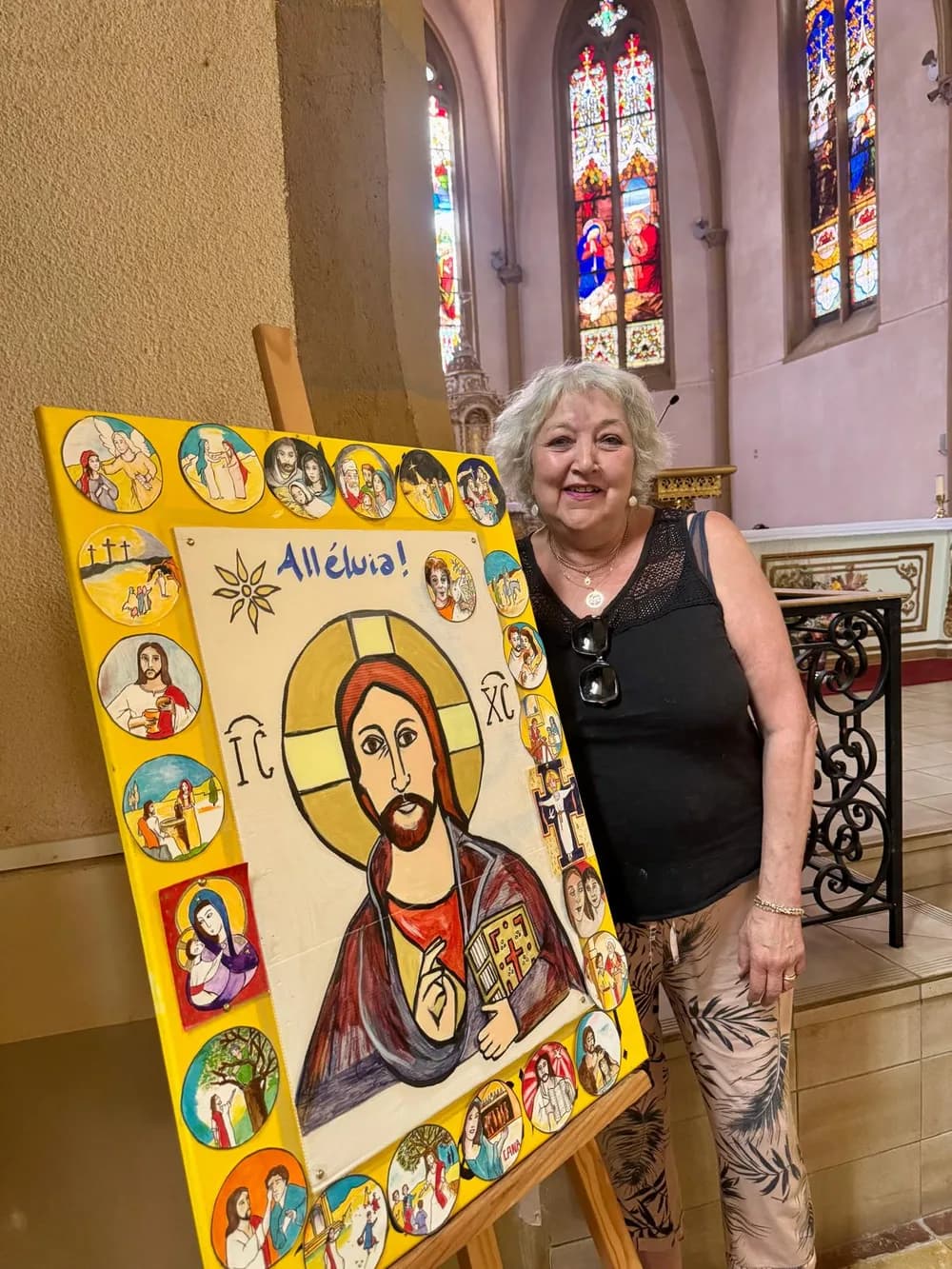Anne-Marie Garandeau, souriante, dans une église aux vitraux colorés, à côté d'un grand tableau aux couleurs vives représentant le Christ, entouré de médaillons peints.