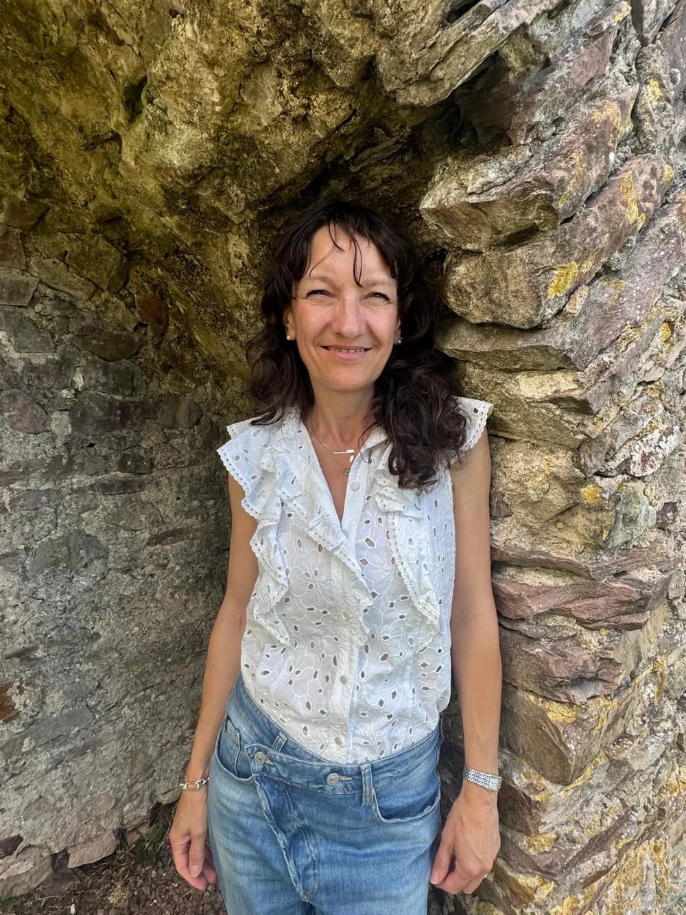 Frédérique Wehr, souriante, en chemisier blanc, adossée à un mur de pierres anciennes.