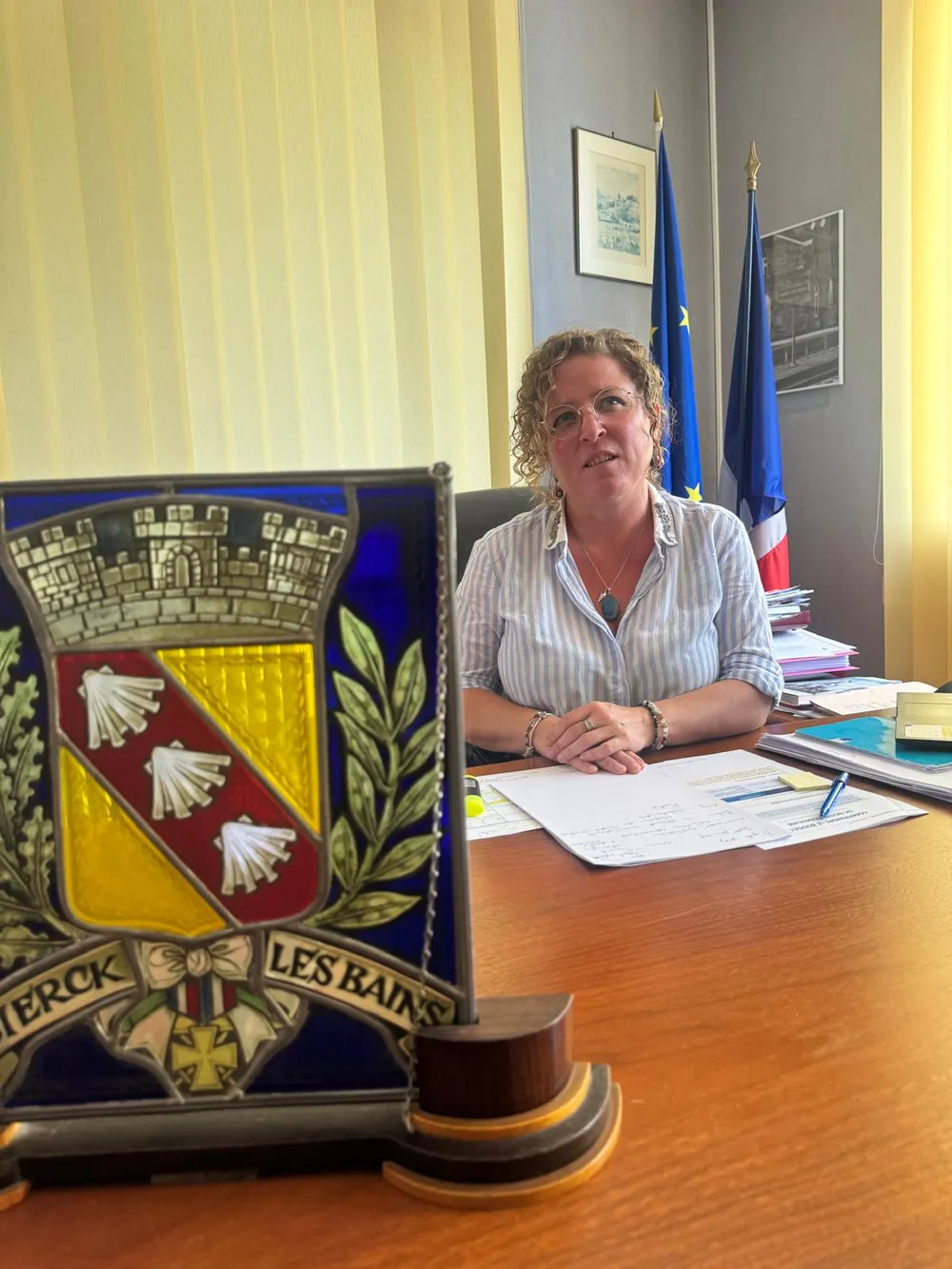 Helen Hammond à son bureau de la mairie, devant les drapeaux européen et français, avec au premier plan un vitrail aux armoiries de Sierck-les-Bains.