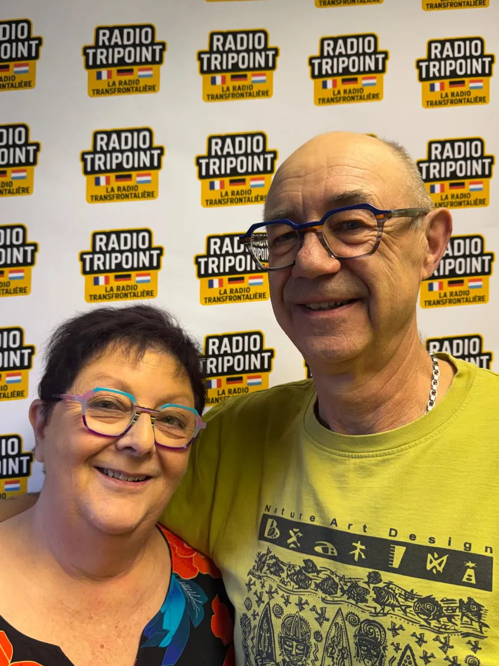 Deux représentants de l'association Une Rose Un Espoir de Sierck-les-Bains, souriants, devant le mur aux logos de Radio Tripoint.