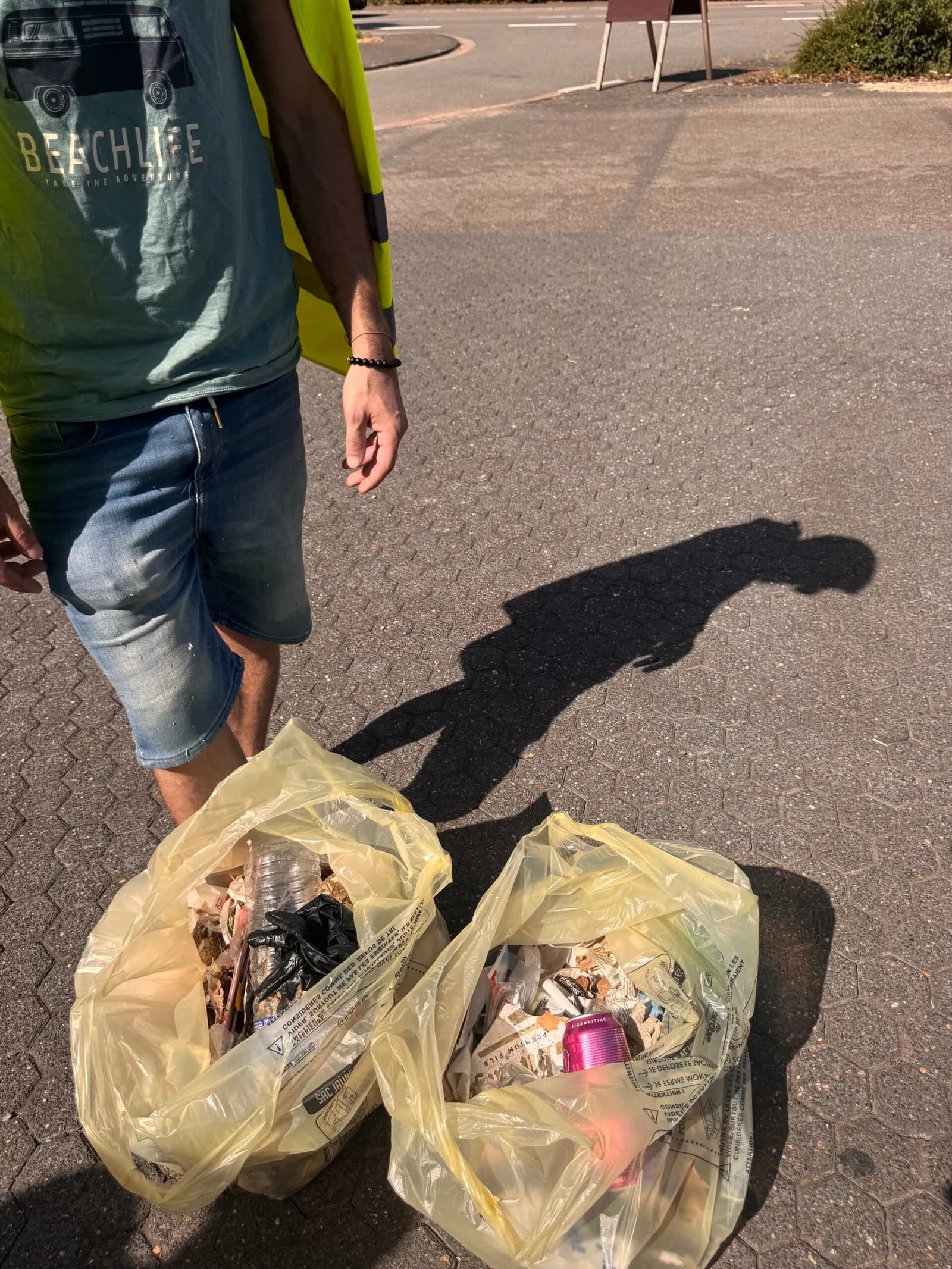 Deux sacs poubelle transparents remplis de déchets posés sur un sol goudronné, près des jambes d'une personne.